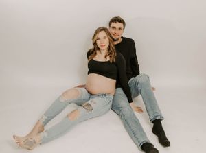 A maternity photoshoot featuring a couple in casual denim and black attire against a light backdrop.