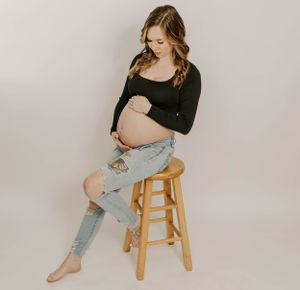 A pregnant woman in ripped jeans sits thoughtfully on a wooden stool during a maternity session.