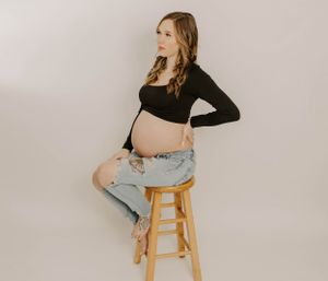 An expectant mother in distressed jeans and black top poses on wooden stool during studio photoshoot.
