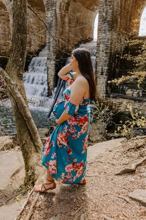 Woman in floral dress stands near waterfall and stone bridge in nature setting.