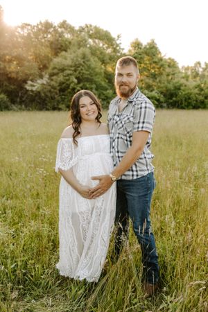 Couple poses in golden field at sunset with woman wearing flowing white lace dress showing baby bump.