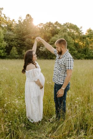 Playful maternity photo of couple dancing in summer field with golden sunlight.