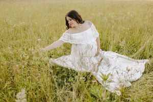 Expecting mother in white lace dress sits gracefully among wildflowers at sunset.