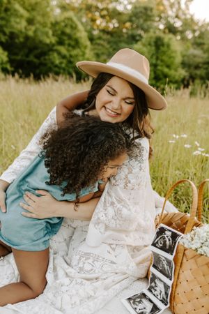 Mother in white dress and hat embraces child on picnic blanket with ultrasound photos nearby.