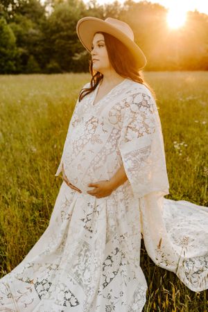 Pregnant woman in flowing lace dress and hat poses in sunlit meadow at golden hour.