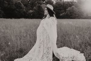 Black and white maternity portrait of woman in lace dress standing in field.
