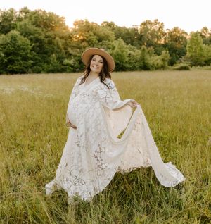 A maternity photoshoot in a golden field at sunset features an elegant white lace dress and wide-brimmed hat.