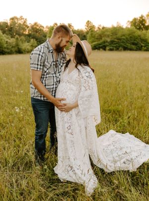 A tender maternity moment captured in a sunlit meadow with a white lace dress and plaid shirt.