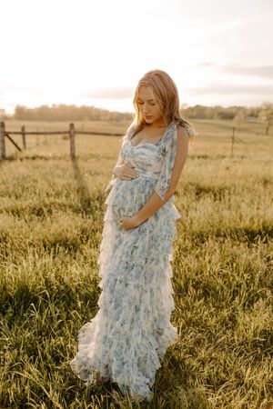 Pregnant woman in flowing blue floral dress standing in golden field at sunset.