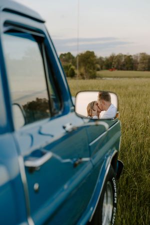 Reflection of couple in side mirror of classic blue truck parked in field.