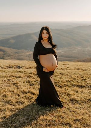 Maternity photo of expectant mother in black dress at sunset on hilltop.