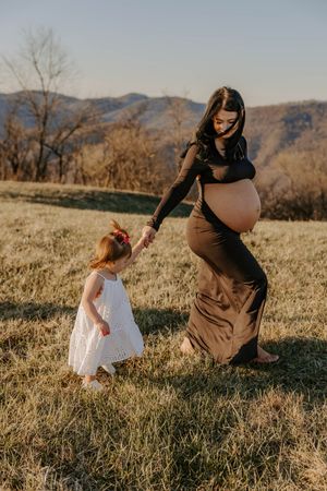 Expecting mother in black dress holds hands with small child on grassy hillside.