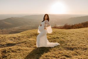 Expectant mother in white gown poses elegantly on hilltop at golden hour.