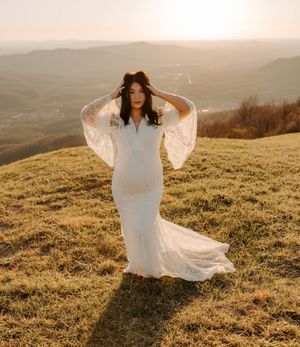 A woman in a flowing white wedding dress poses on a hilltop during golden hour with mountains in the background.
