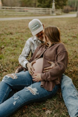 A couple sits together in a grassy field during a maternity photoshoot wearing casual denim and sweaters.