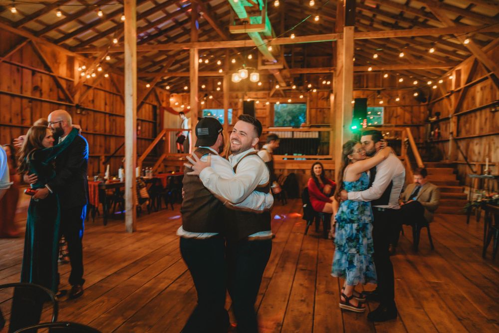 Couples dancing together on a rustic wooden dance floor inside a warmly lit barn with string lights overhead.
