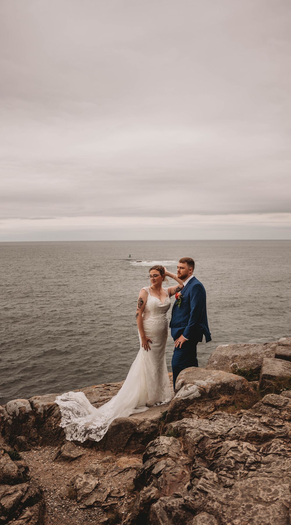 Newlywed couple stands on rocky coastal cliff overlooking misty ocean on their wedding day.