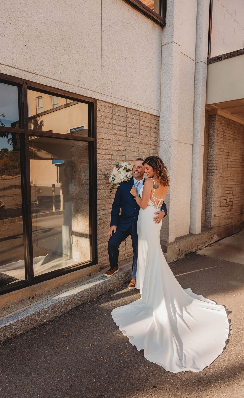 A couple in formal attire stands beside a reflective glass window as sunset light illuminates the bride's flowing white gown.