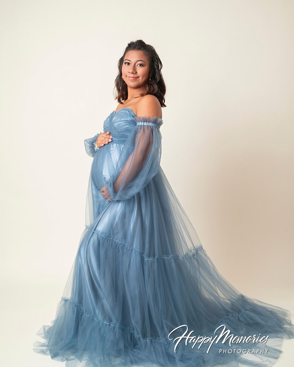 Pregnant woman in elegant off-shoulder blue tulle gown posing for maternity photography session.