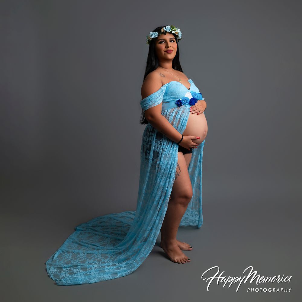 Pregnant woman in blue lace maternity gown with floral crown poses elegantly for Happy Memories Photography studio portrait.