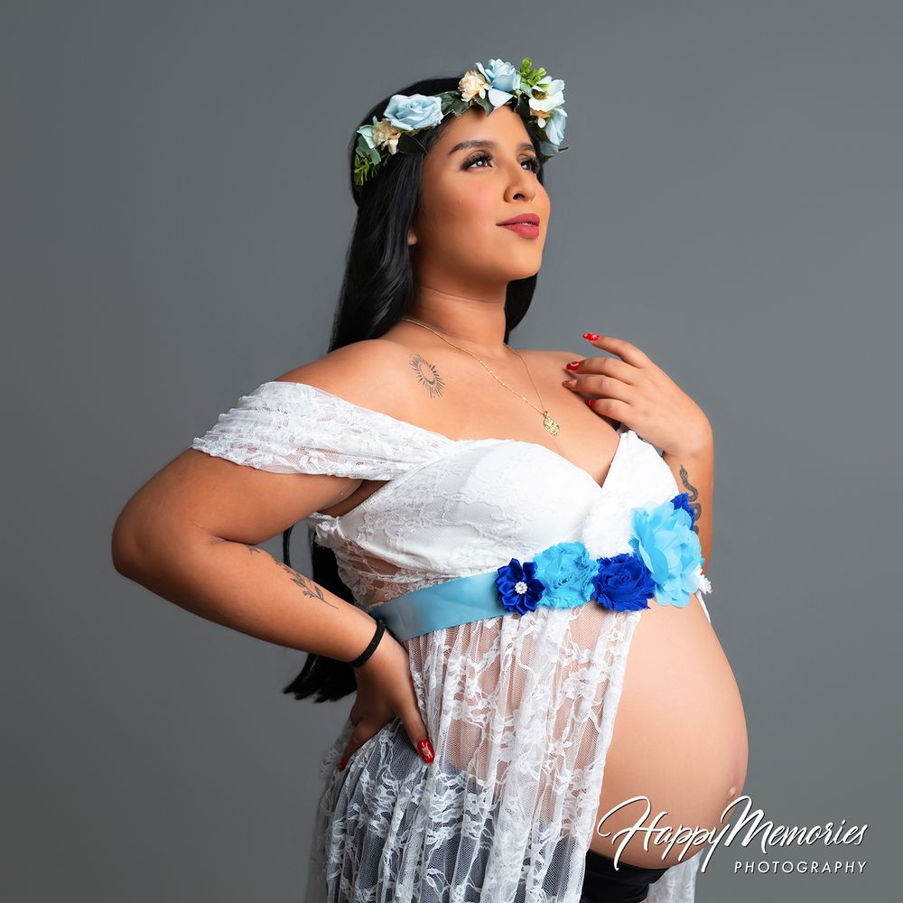 Pregnant woman in white lace off-shoulder gown with blue floral sash and flower crown, maternity photography studio portrait.