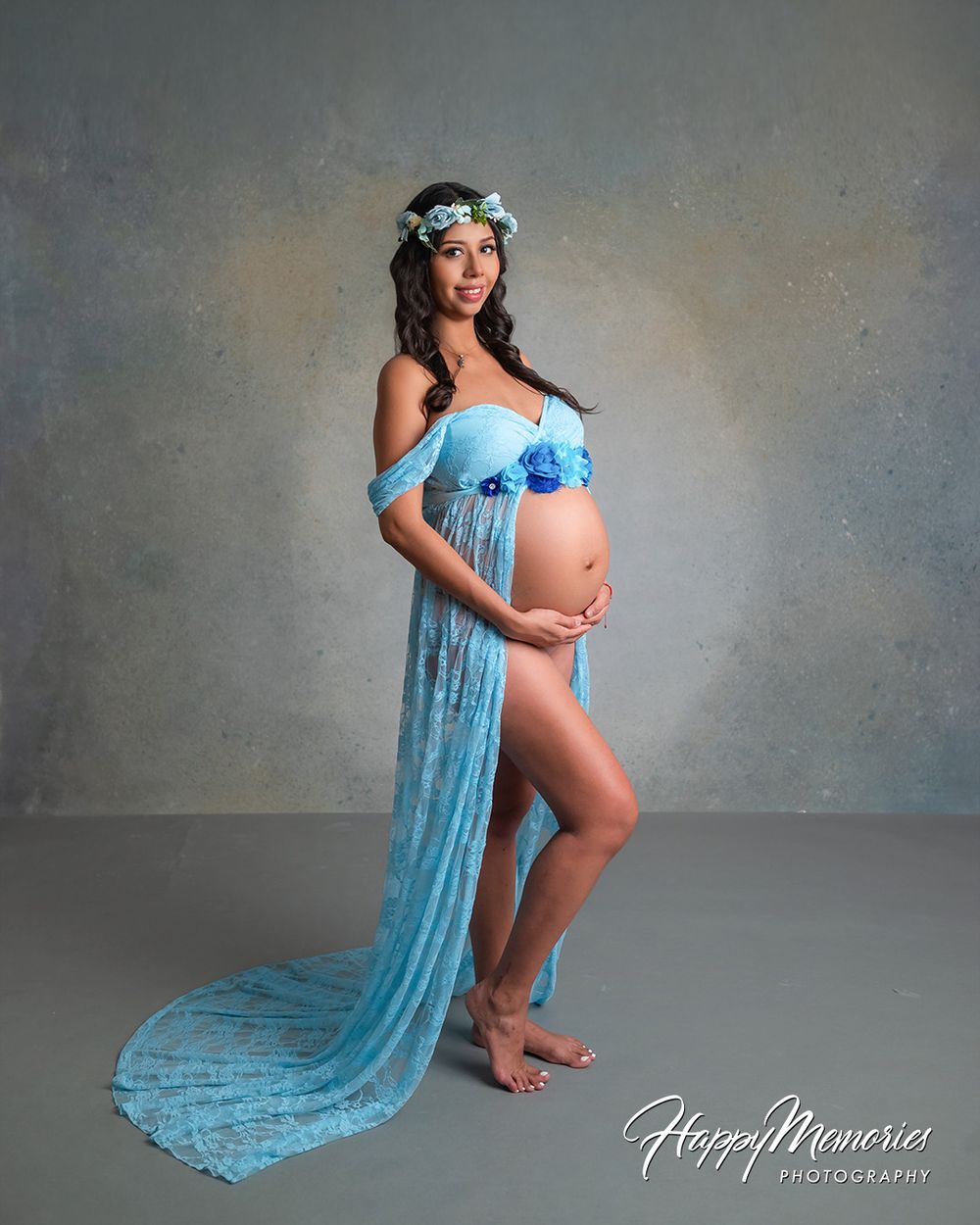 Pregnant woman in blue lace maternity gown with floral crown poses elegantly in professional studio photography session.