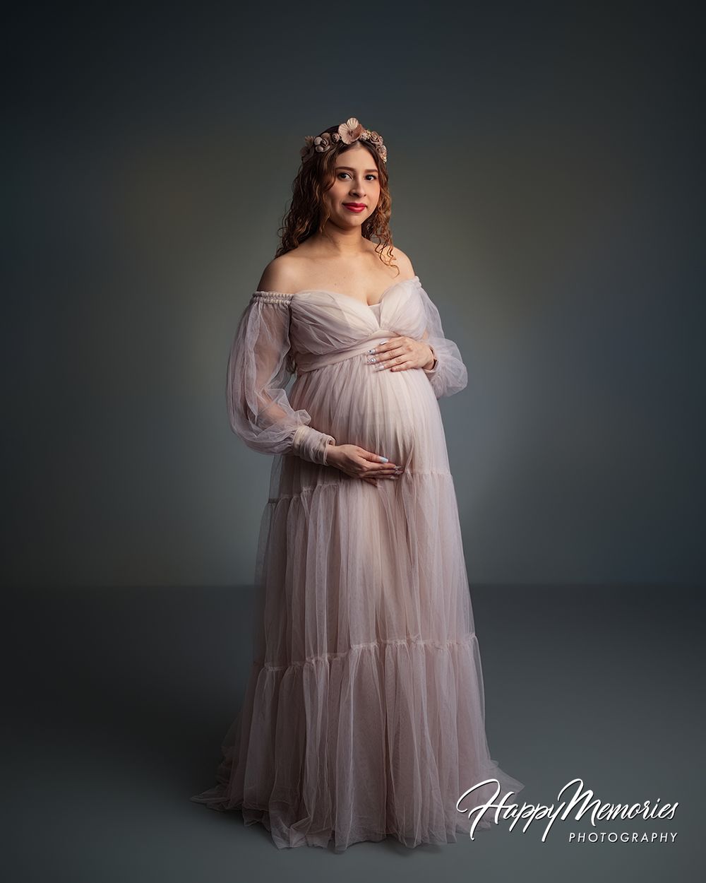 Pregnant woman in elegant blush tulle off-shoulder maternity gown with floral crown poses gracefully in studio.