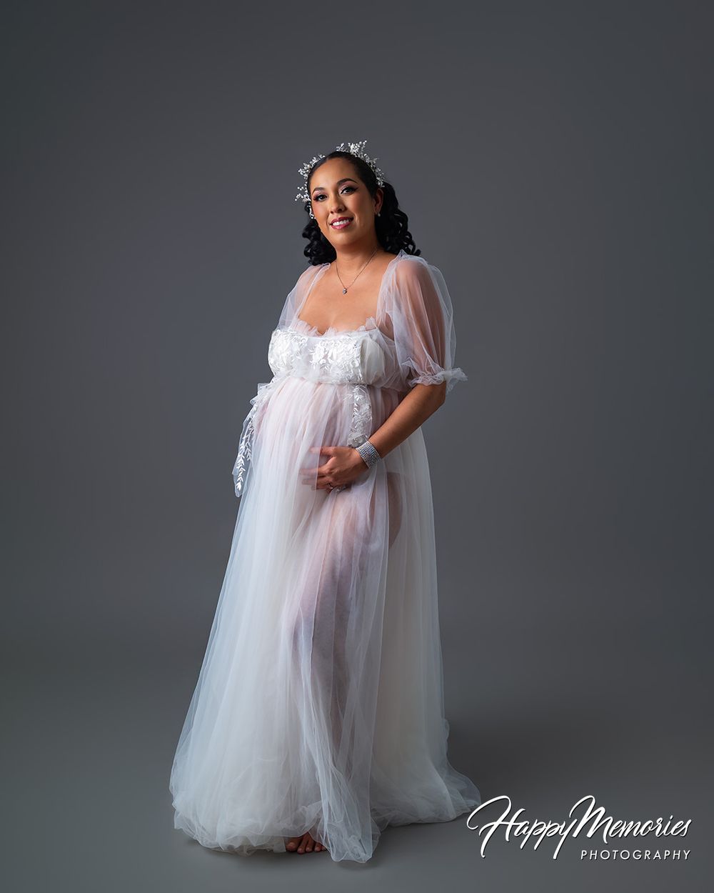 Pregnant woman in white tulle maternity gown with floral crown poses gracefully in professional studio photography session.