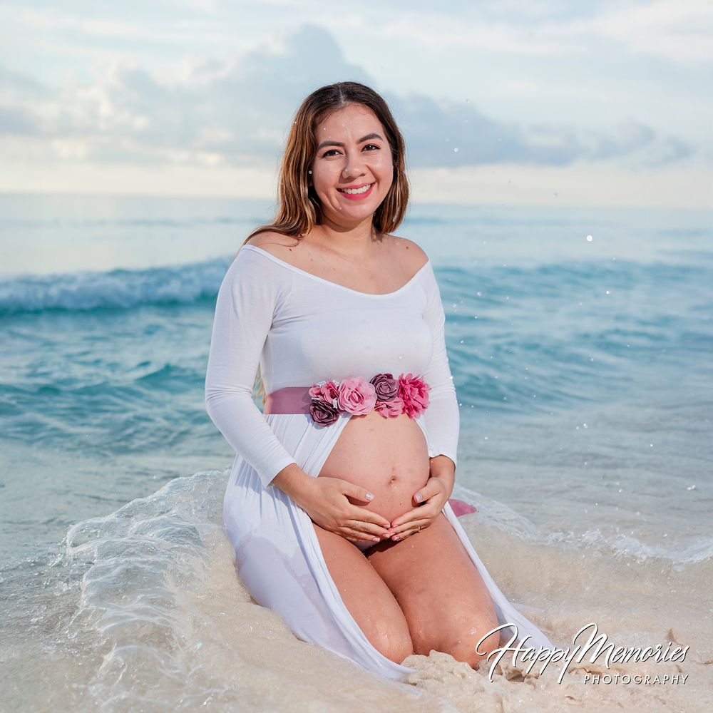 Pregnant woman in white off-shoulder dress with floral belt kneeling on beach shore, smiling at camera.