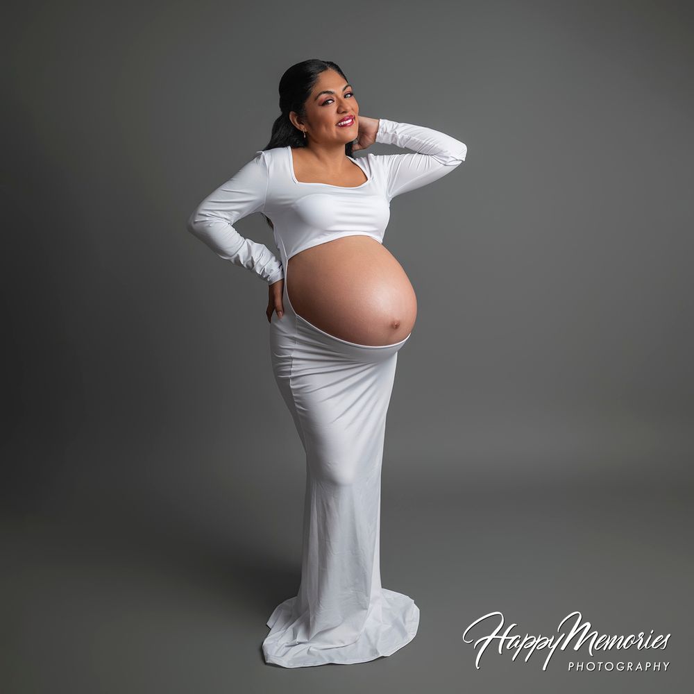 Pregnant woman in white maternity gown posing for a professional studio photo shoot by Happy Memories Photography.