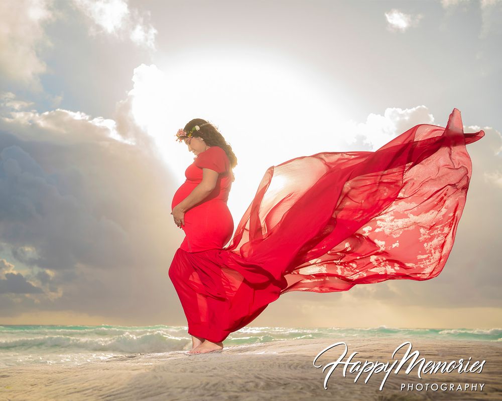 Pregnant woman in flowing red gown on beach with dramatic sky, maternity photography by Happy Memories.