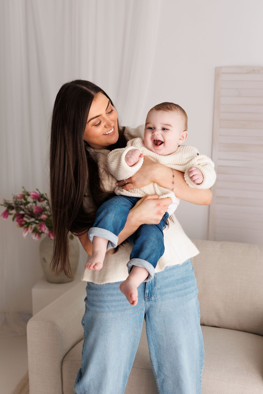 Happy young mother holding laughing baby in white knit outfit in bright living room.