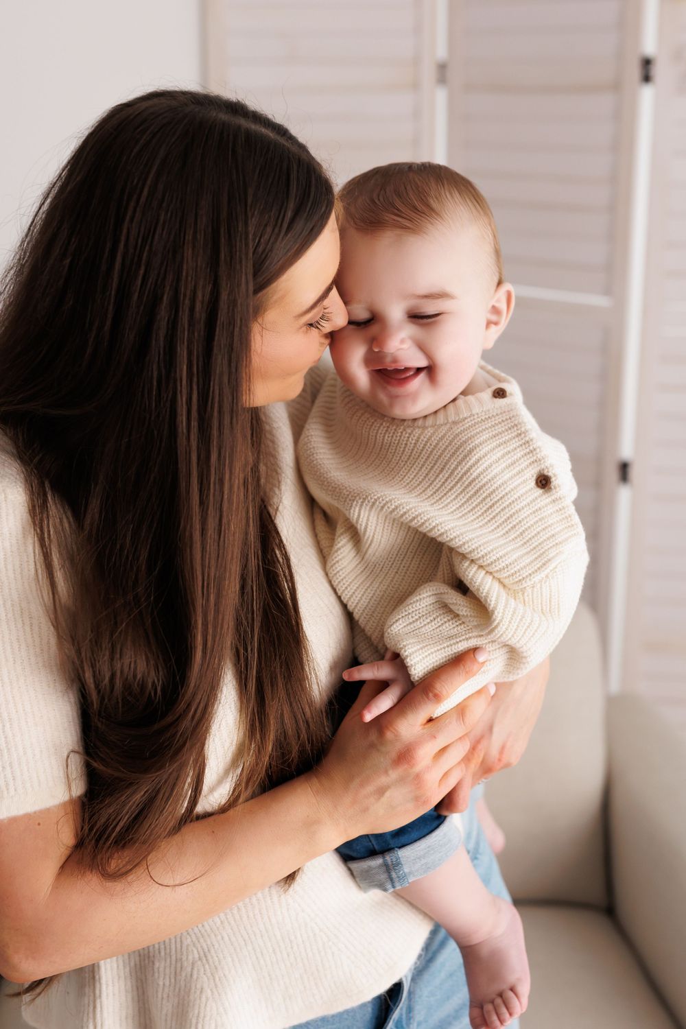 A loving mother with long dark hair holds her laughing baby dressed in a cream knit sweater indoors.