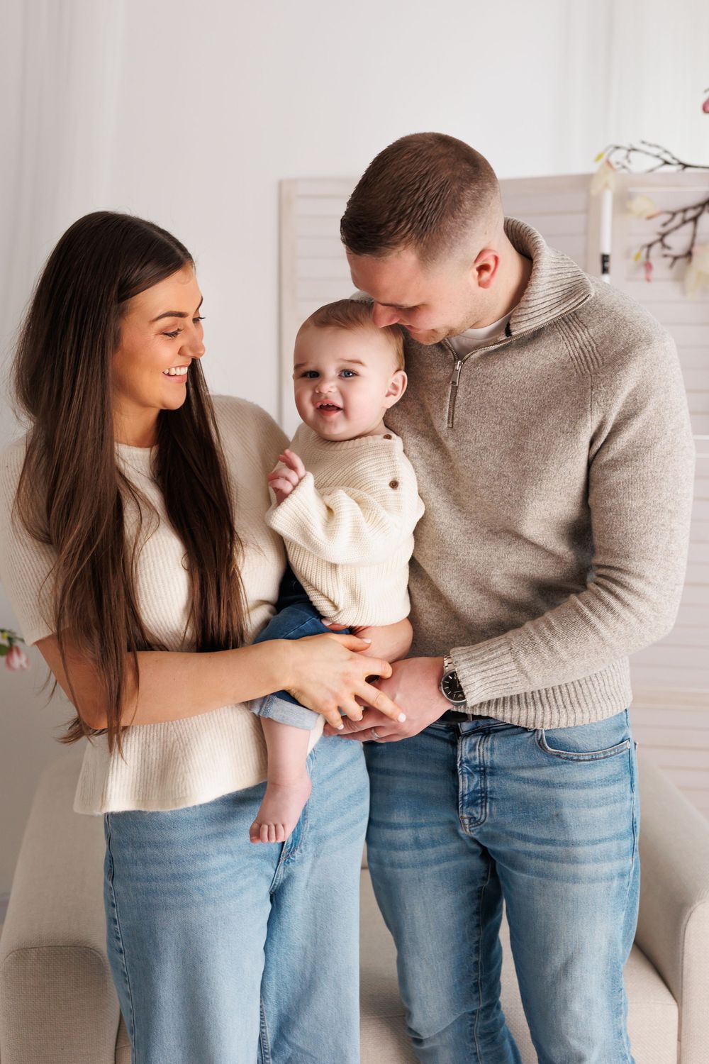 Young family with mother, father, and baby in white sweater posing together in bright white room.