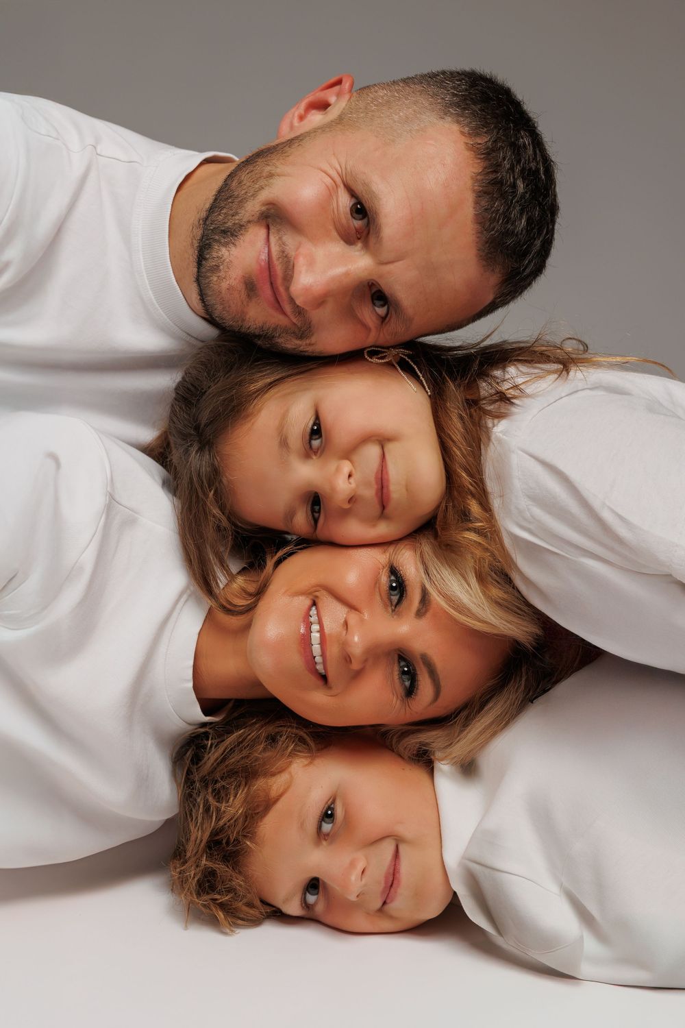Happy family of four lying together stacking heads smiling at camera wearing white tops on gray background.
