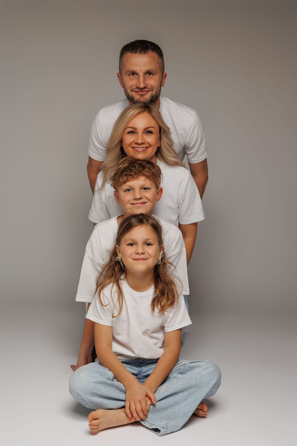 Family of four in matching white t-shirts posing in a stacked portrait formation against a gray background.