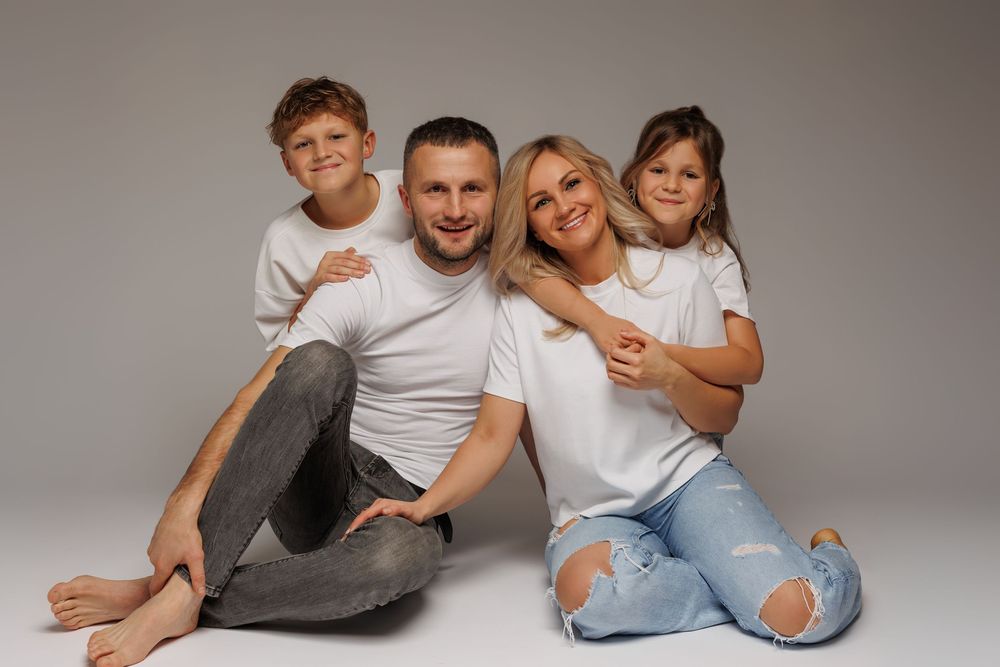 Happy family of four in white shirts sitting together on gray background, smiling parents with two children.