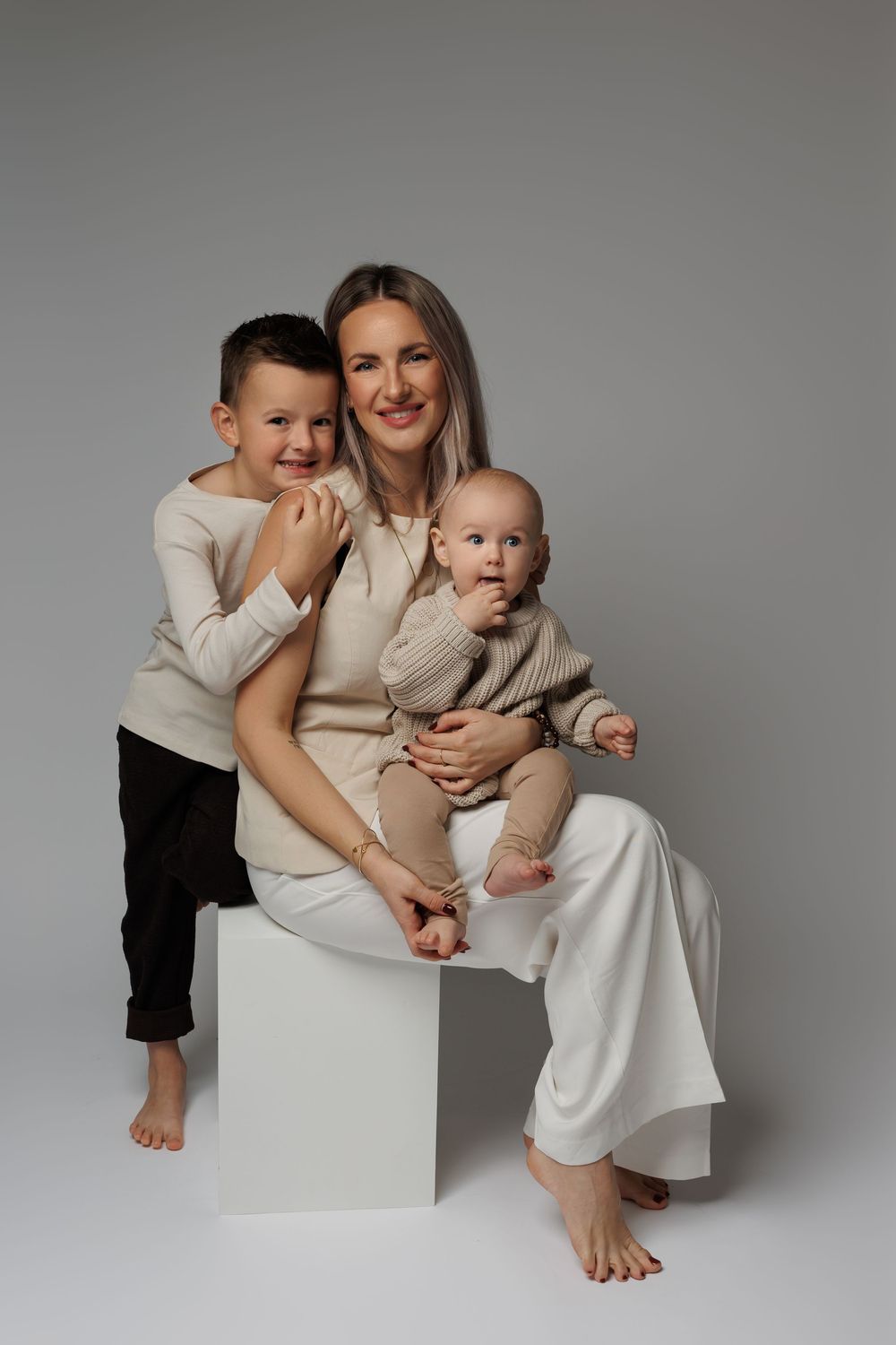 A smiling mother sits on a white cube holding a baby, with her young son standing beside her, all in neutral tones.
