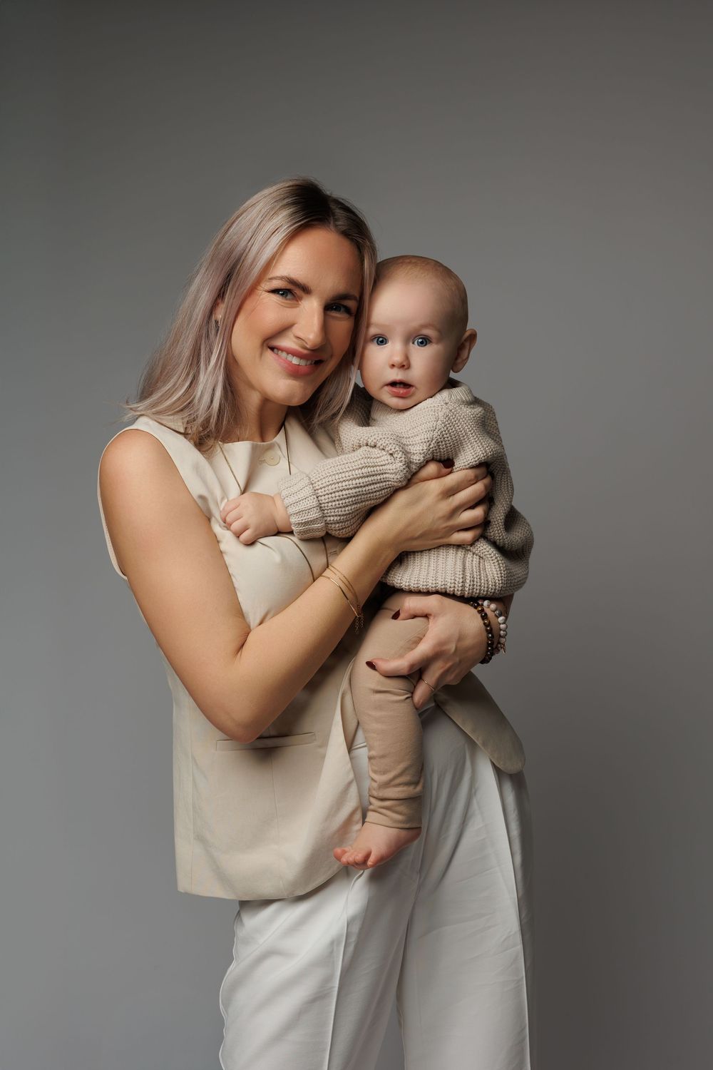 Smiling blonde mother in beige outfit holding her blue-eyed baby dressed in a knit sweater against a gray background.