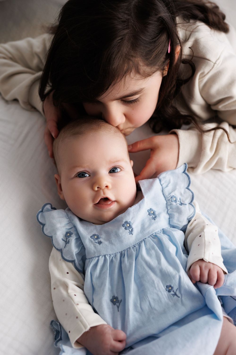 Young girl in beige top kisses baby sister wearing a blue floral dress while lying on a white bed.
