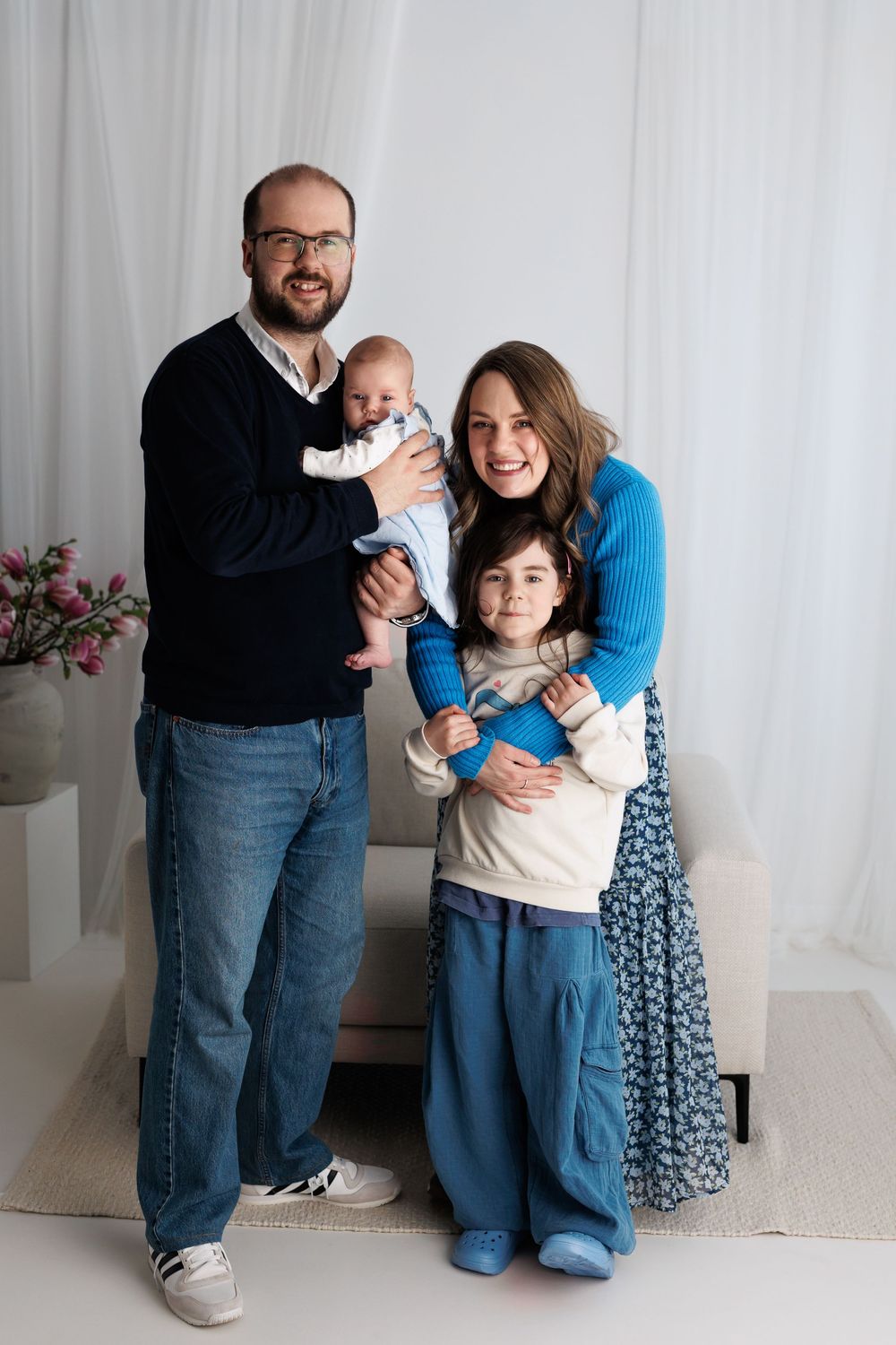 Happy family of four posing for a portrait, parents holding two young children, dressed in blue tones against white backdrop.