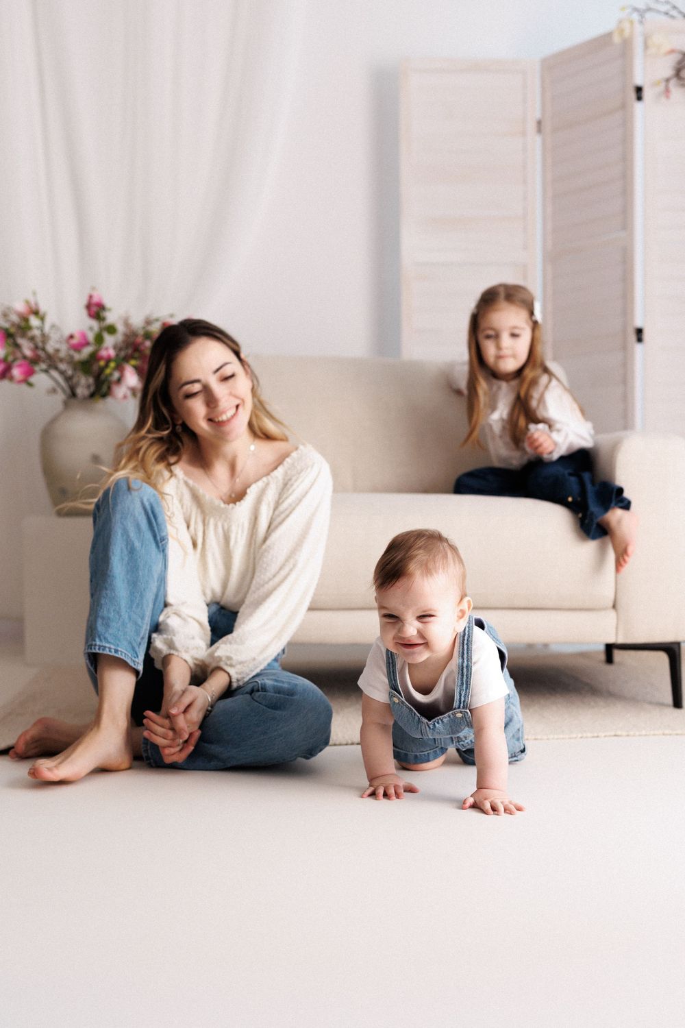 Happy mother sitting on floor with crawling baby while older daughter plays on sofa in bright living room.