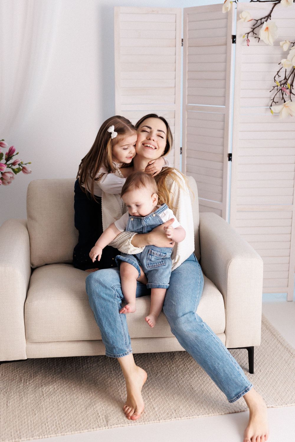 Happy mother sitting on sofa hugging two children, older daughter and baby in denim overalls, in bright living room.