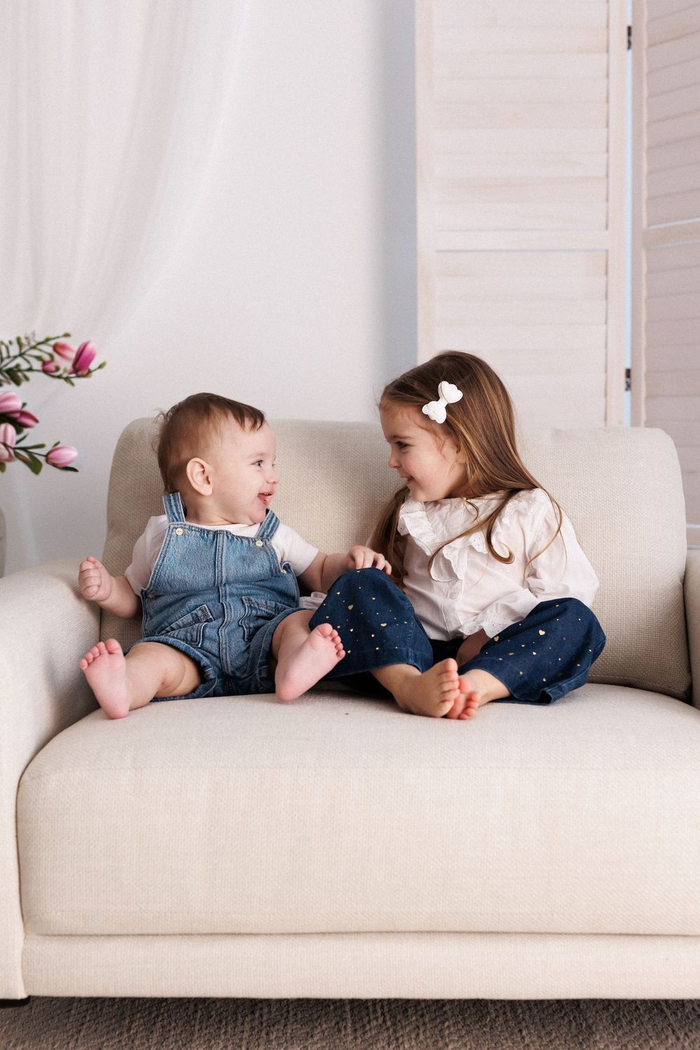 Two young siblings sitting barefoot on a beige sofa, smiling and interacting in a bright white living room.