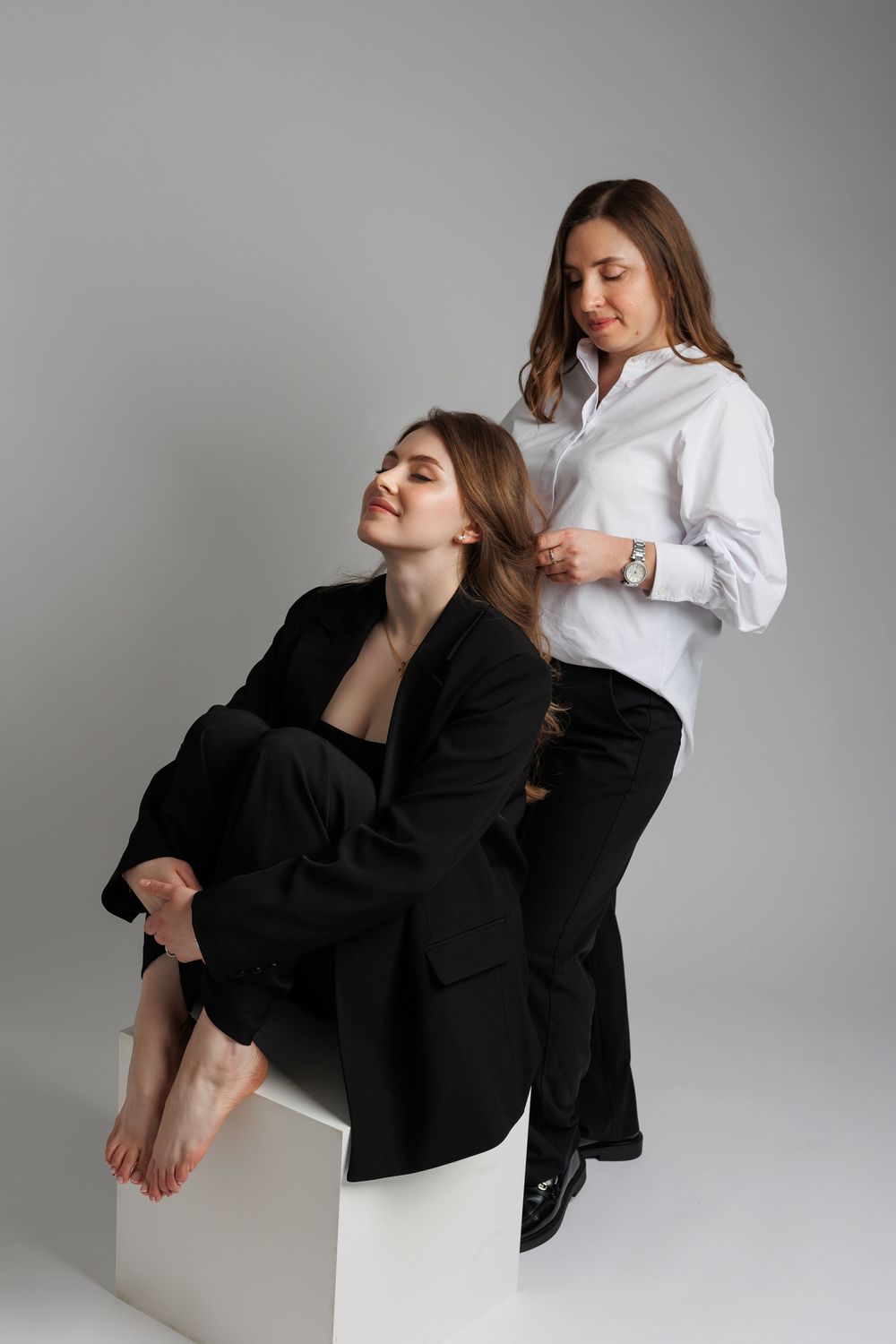 Two women posing in a studio, one seated barefoot in a black blazer, the other standing in a white shirt styling her hair.