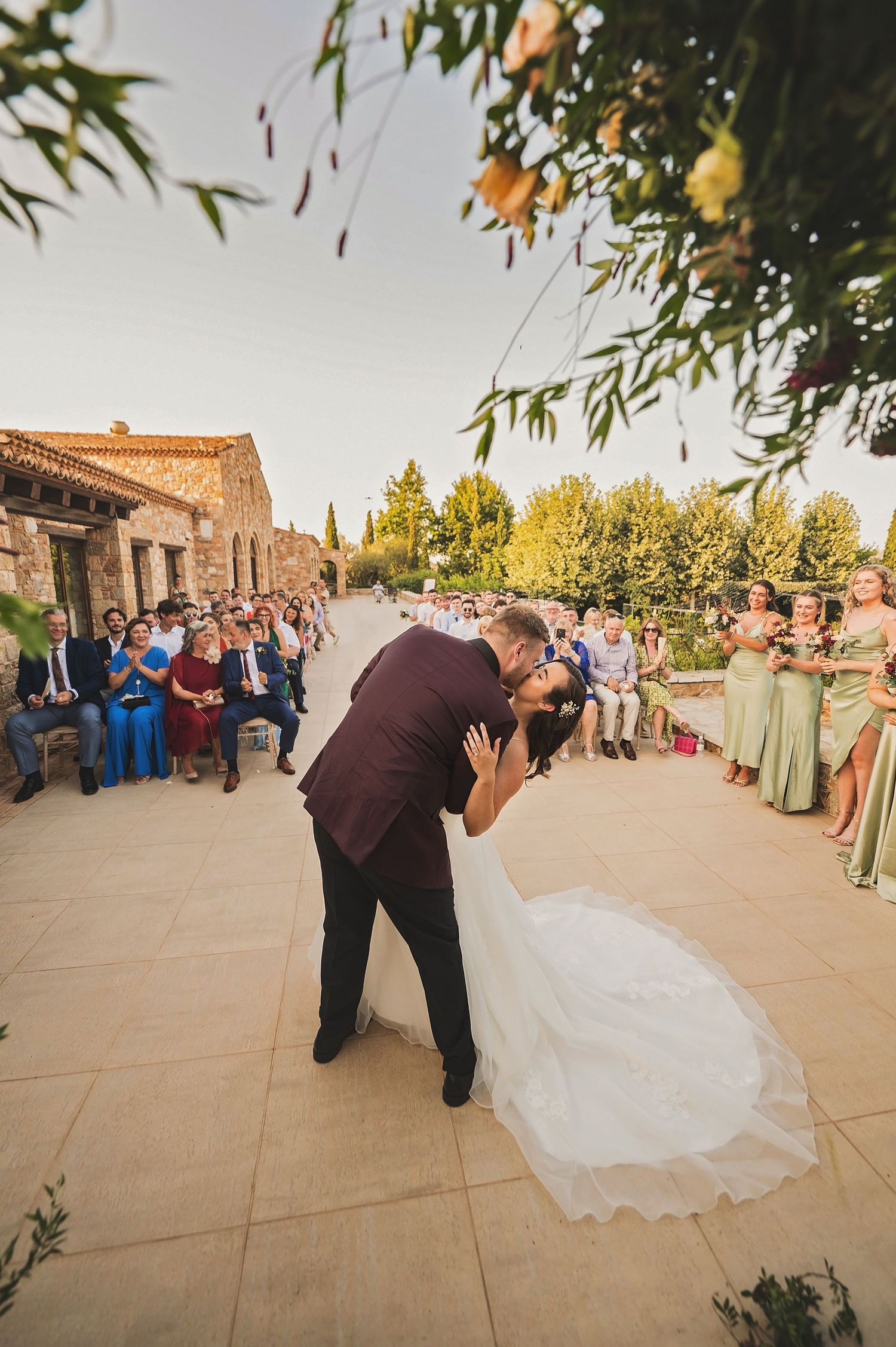 A romantic wedding kiss during the ceremony at a rustic Mediterranean venue with guests and bridesmaids in attendance.