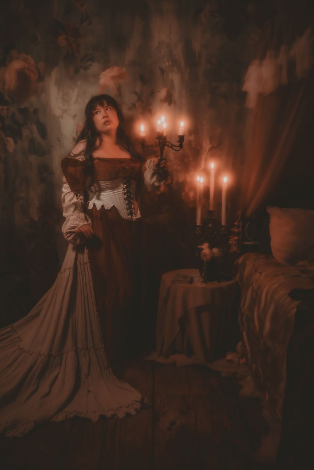 A woman in a Victorian-era gown stands by candlelight in a dark, moody room with dramatic shadows.