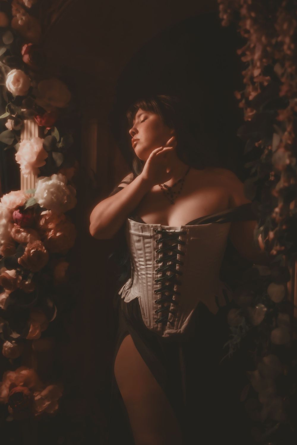 Woman in white corset poses gracefully among lush floral arrangements in moody, romantic lighting.