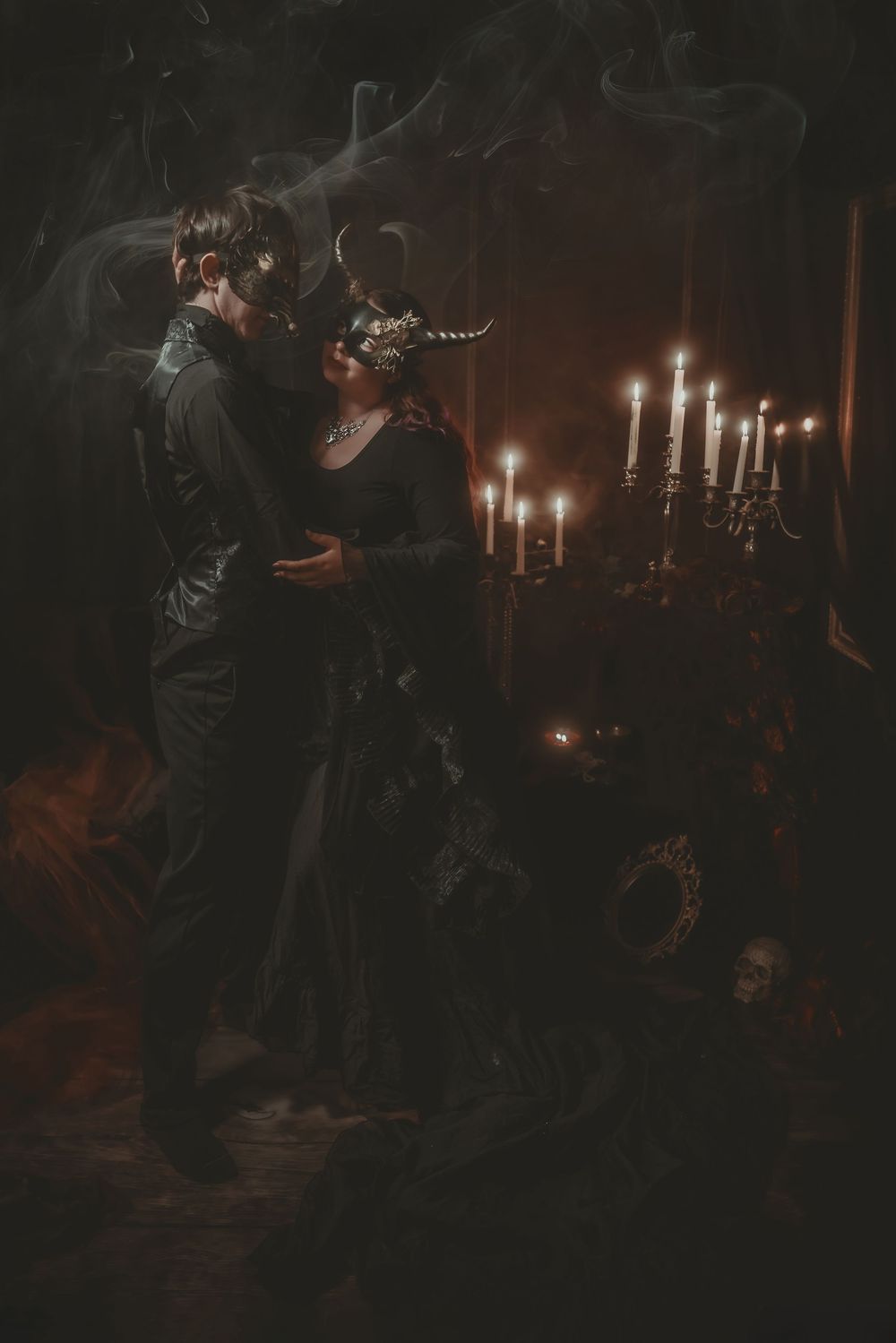 A couple in black Gothic attire with masks embrace by candlelight in a dark, moody, atmospheric setting.