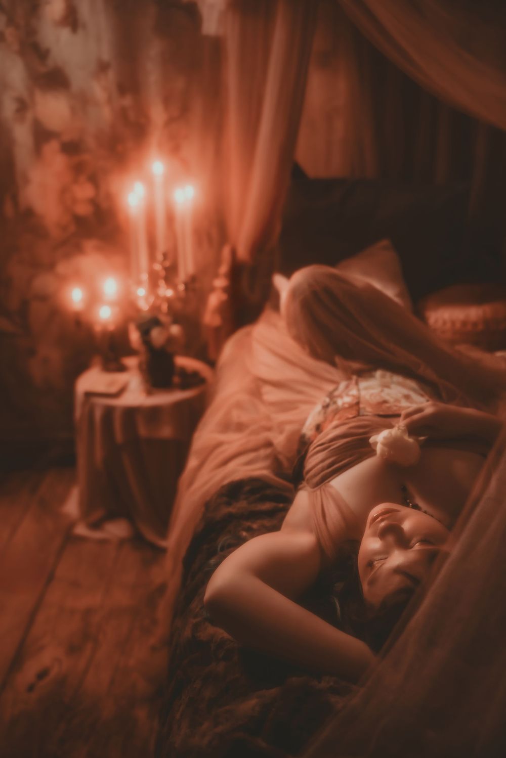 Woman reclining on a bed in warm candlelight, surrounded by sheer drapes in a romantic, moody setting.